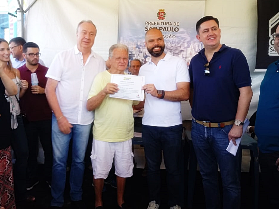 Foto do prefeito Bruno Covas entregando o certificado de reconhecimento ao morador Sr. Antonio Giacuinto. Estão na foto, além do prefeito e do Sr. Antonio, o subprefeito e uma outra pessoa.